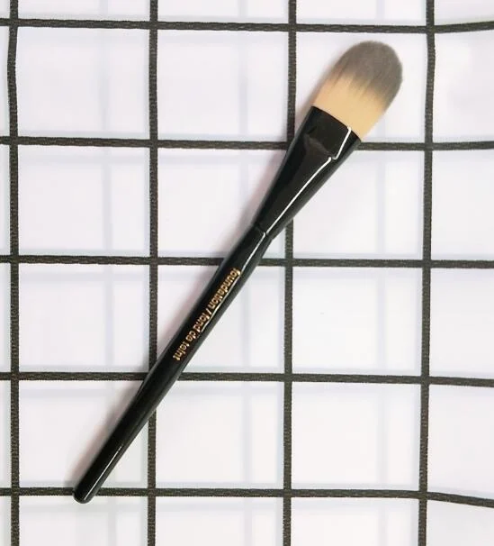 A black-handled tongue-shaped foundation brush with two-tone bristles lying on a white and black grid background.