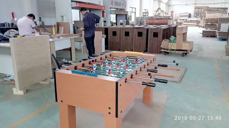 Workers assembling table cabinets with a finished beech coloured soccer table body in the foreground and furth