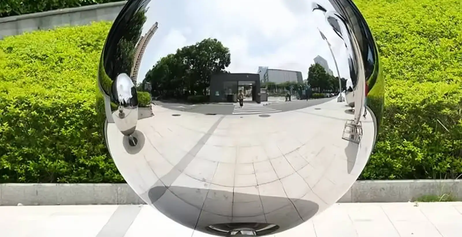 Large mirror polished stainless steel sphere standing on a paved plaza in front of a clipped hedge