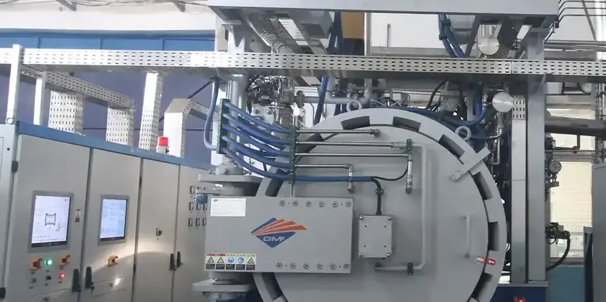 Industrial vacuum heat-treatment furnace with round front door next to electrical control cabinets in a factory workshop